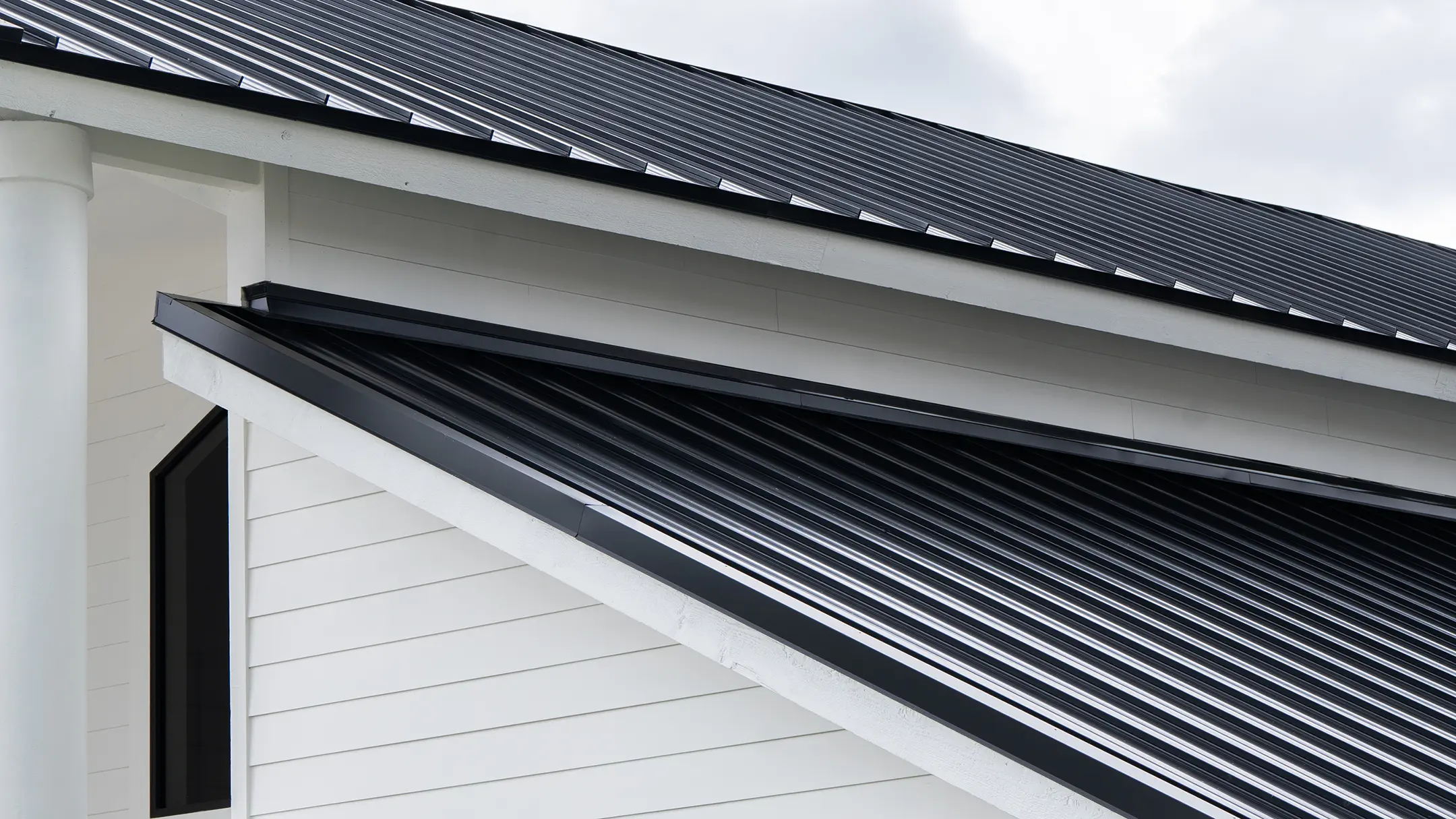 Close-up view of a modern building's exterior, featuring white horizontal siding and a black metal roof with clean, sharp angles. The image highlights the building's contemporary architectural lines.