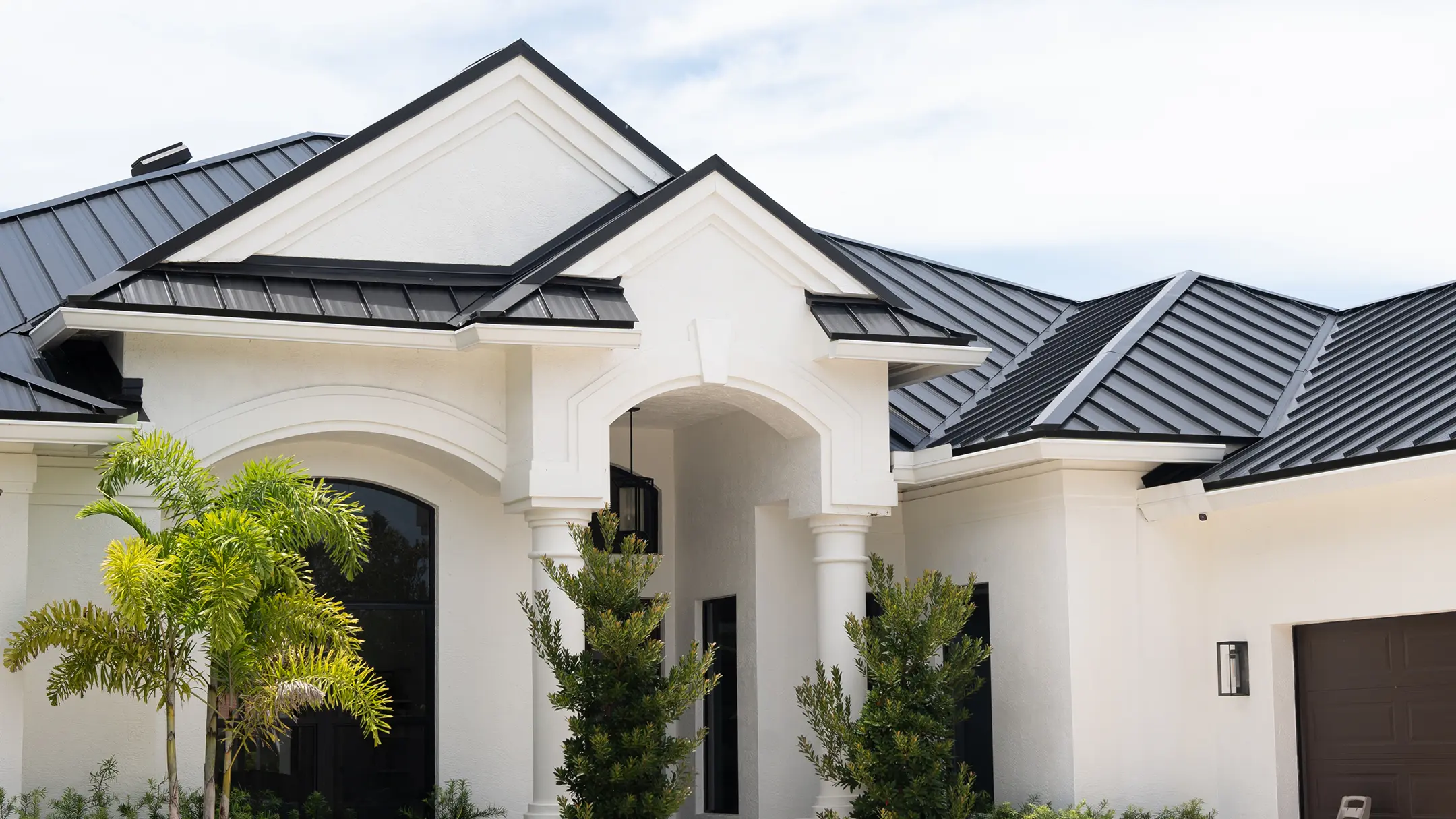 Modern white house with a black metal roof, arched windows, and decorative columns. Green bushes and a palm tree are in front of the entrance under a partly cloudy sky.