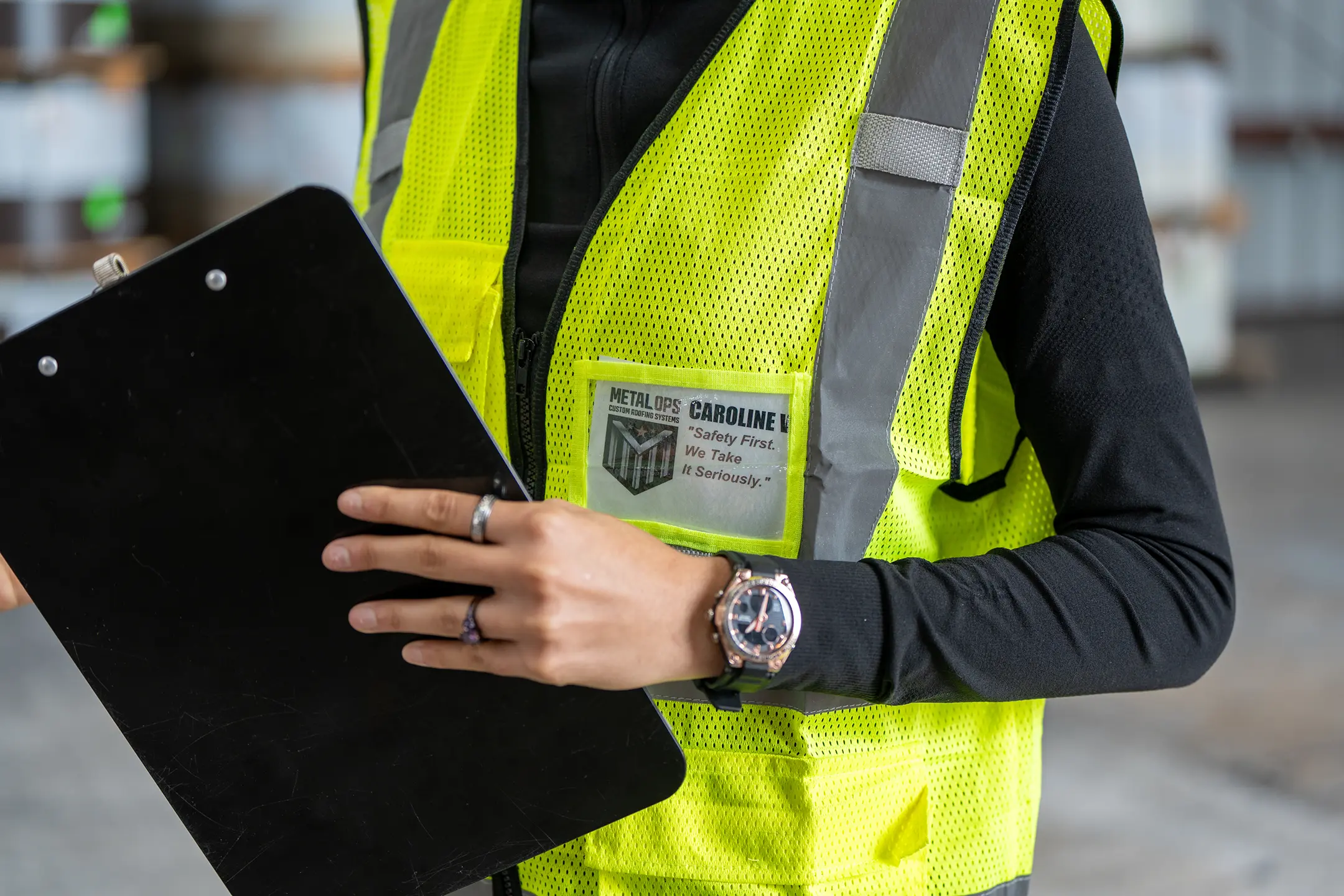 A person in a yellow safety vest holding a clipboard. The vest has a name tag that reads “METAL OPS: CAROLINE,” and the person is wearing rings and a watch. The background appears to be an industrial setting.