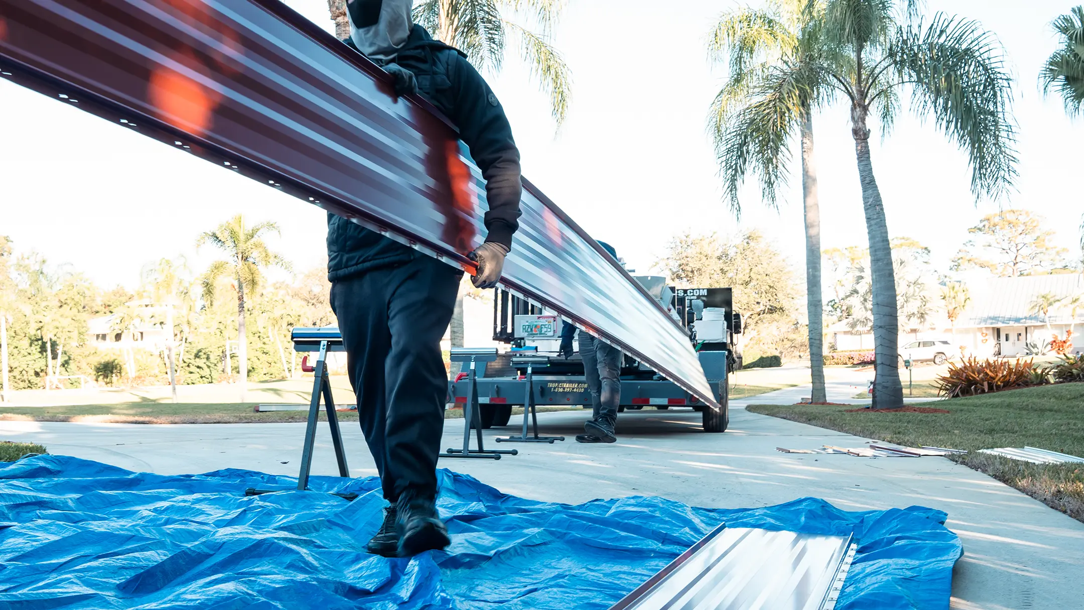 A person in work clothes carries a long, red metal sheet outdoors, standing on a blue tarp, with palm trees and a work truck in the background.