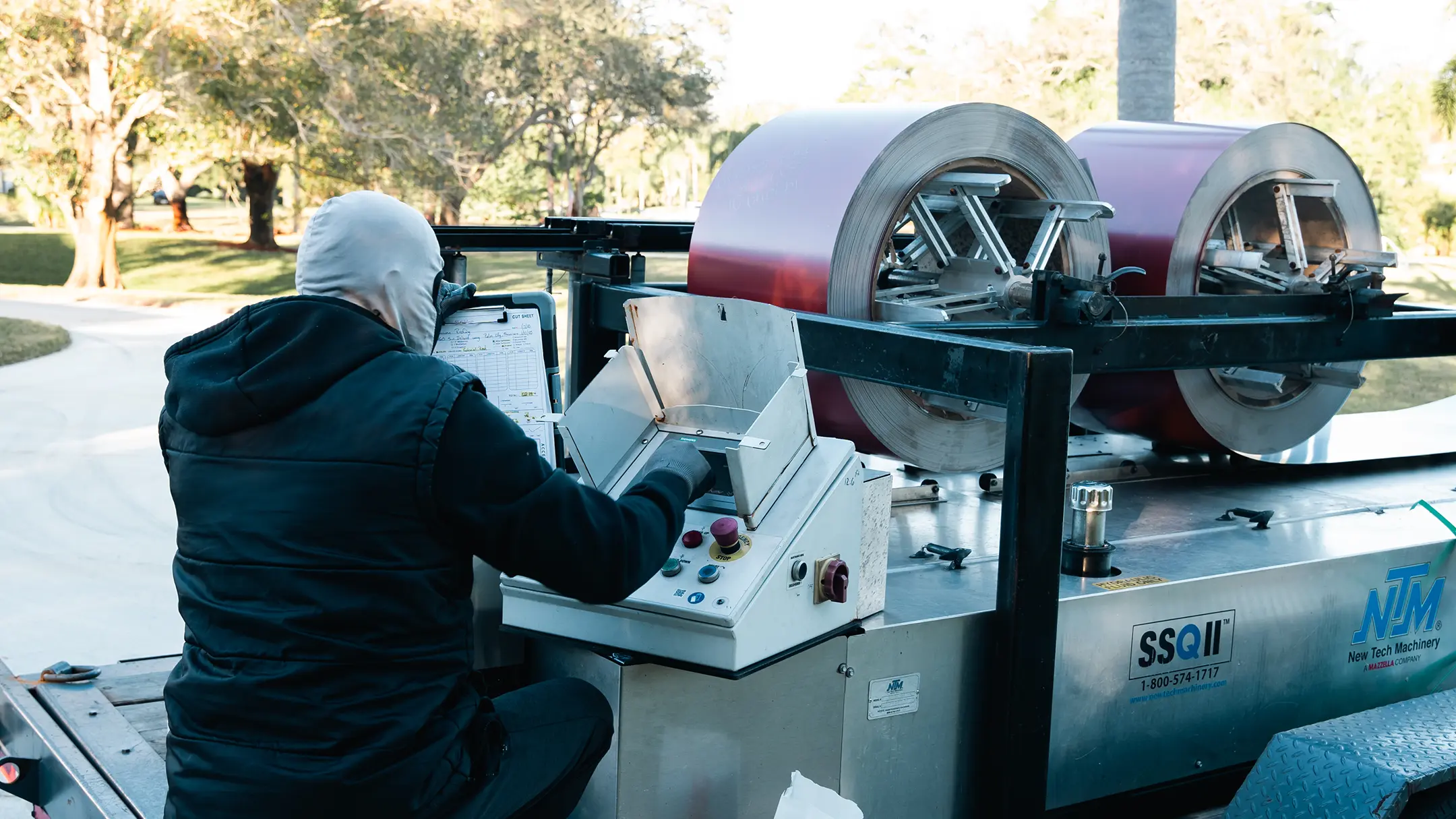 A person in a jacket and hood operates a large industrial machine with metal rolls and a control panel outdoors during the day. Trees and sunlight are visible in the background.