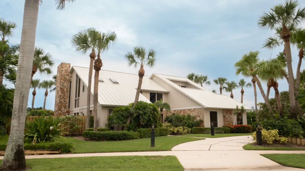 A modern house with a metal roof and stone accents is surrounded by palm trees and lush landscaping under a partly cloudy sky. A curved sidewalk leads to the entrance.