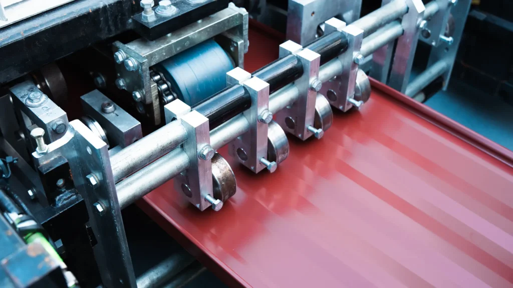 Close-up of an industrial machine rolling and shaping a sheet of red metal, likely in a manufacturing or production facility. The machinery features rollers and metal supports guiding the sheet through the process.