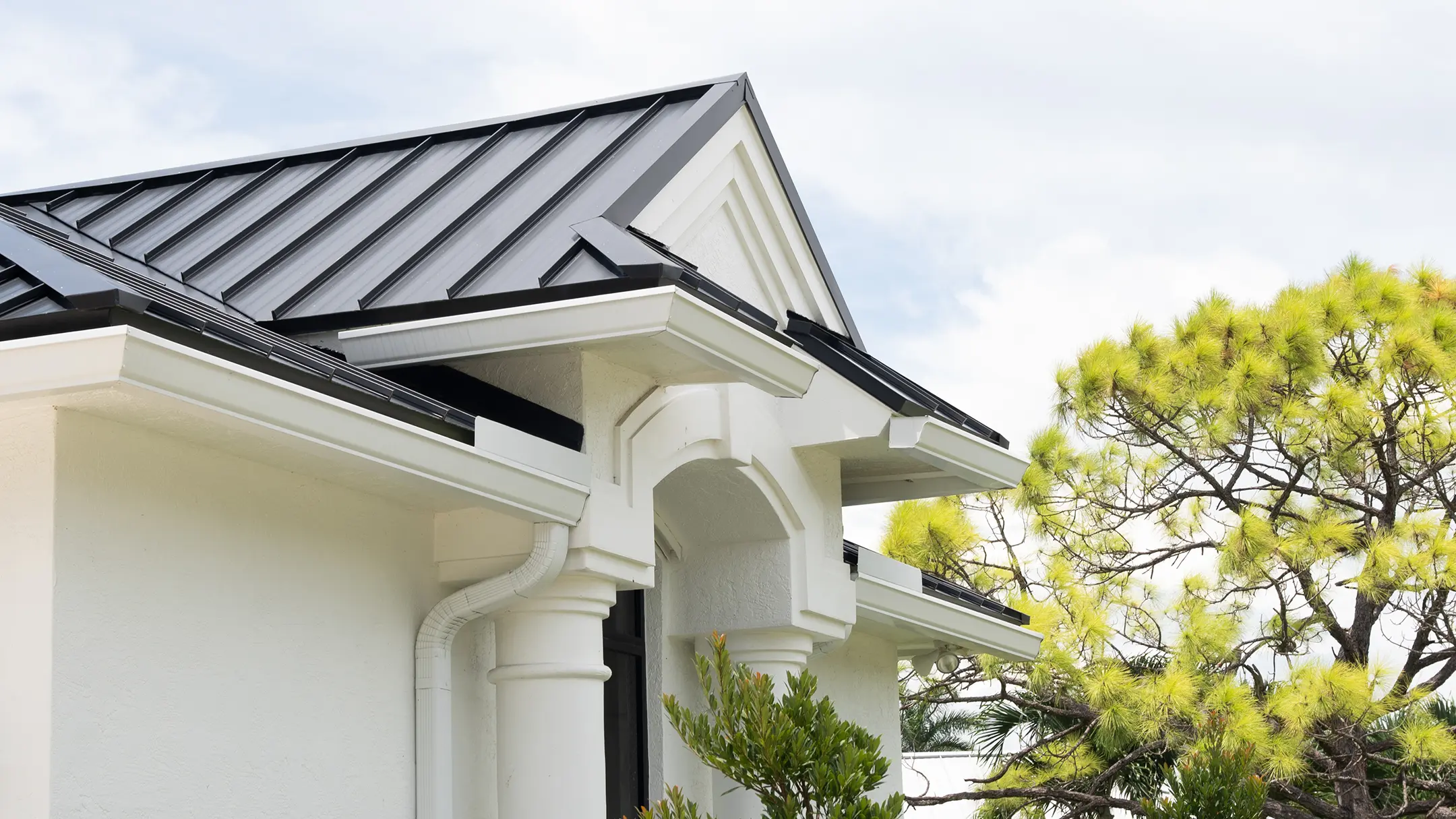 Close-up view of a house with a white exterior and dark metal roof, featuring decorative columns and trim, with green trees and a cloudy sky in the background.