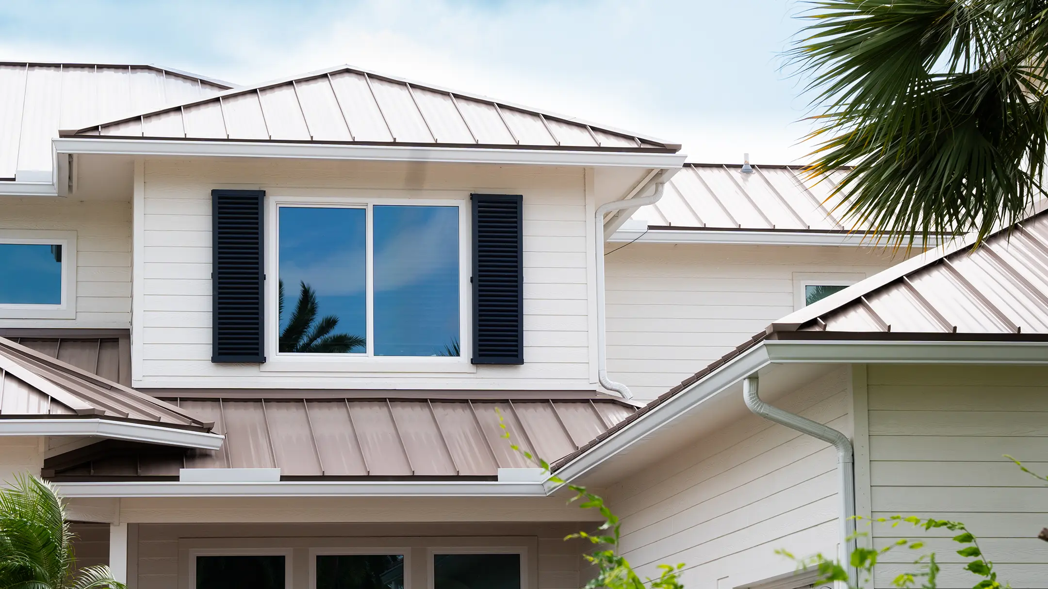 A close-up of a modern house exterior with white siding, brown metal roofing, a double window with black shutters, and a palm tree partially visible on the right side.
