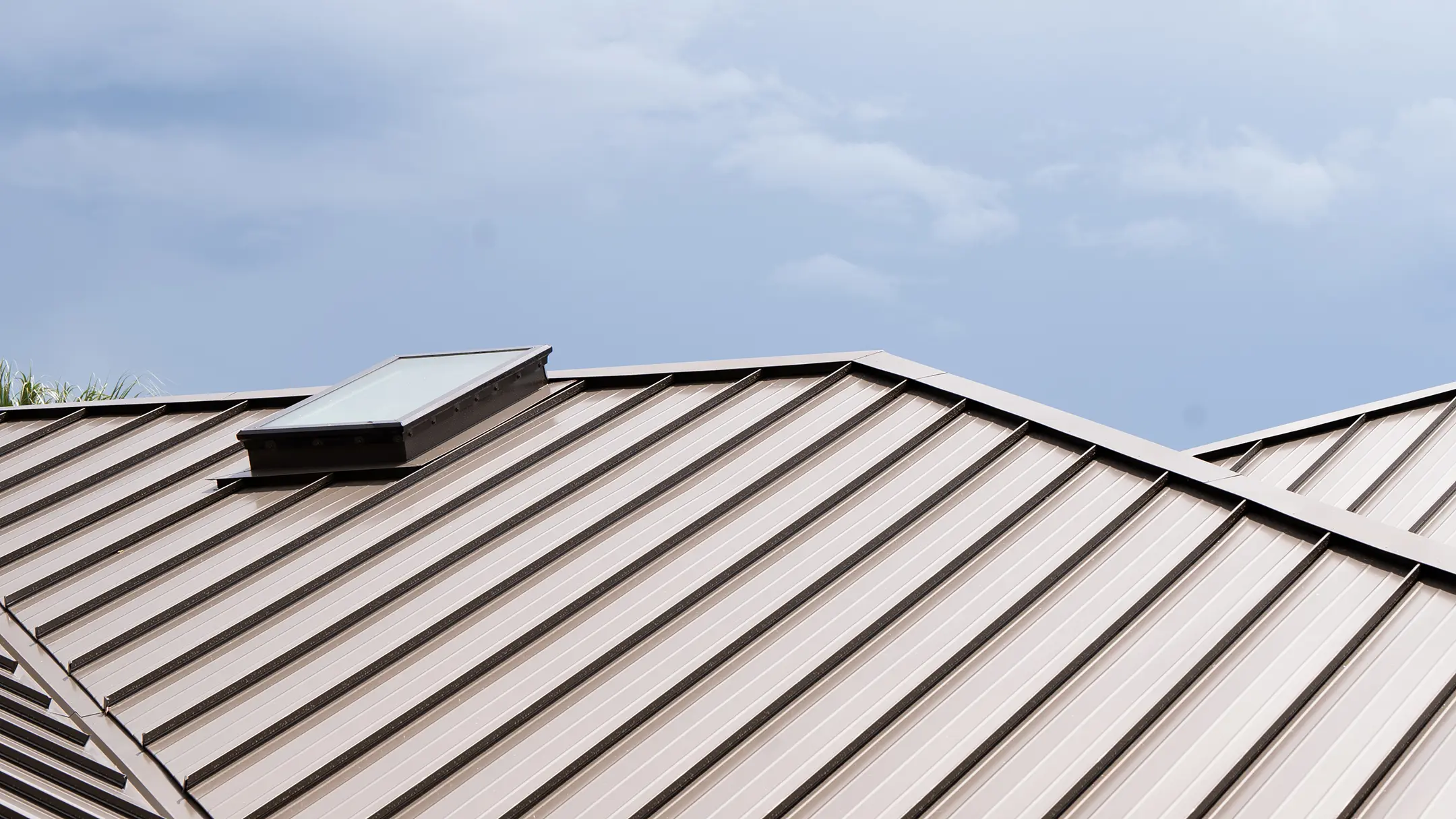 A close-up view of a light-colored metal roof with a rectangular skylight, set against a blue sky with light clouds.
