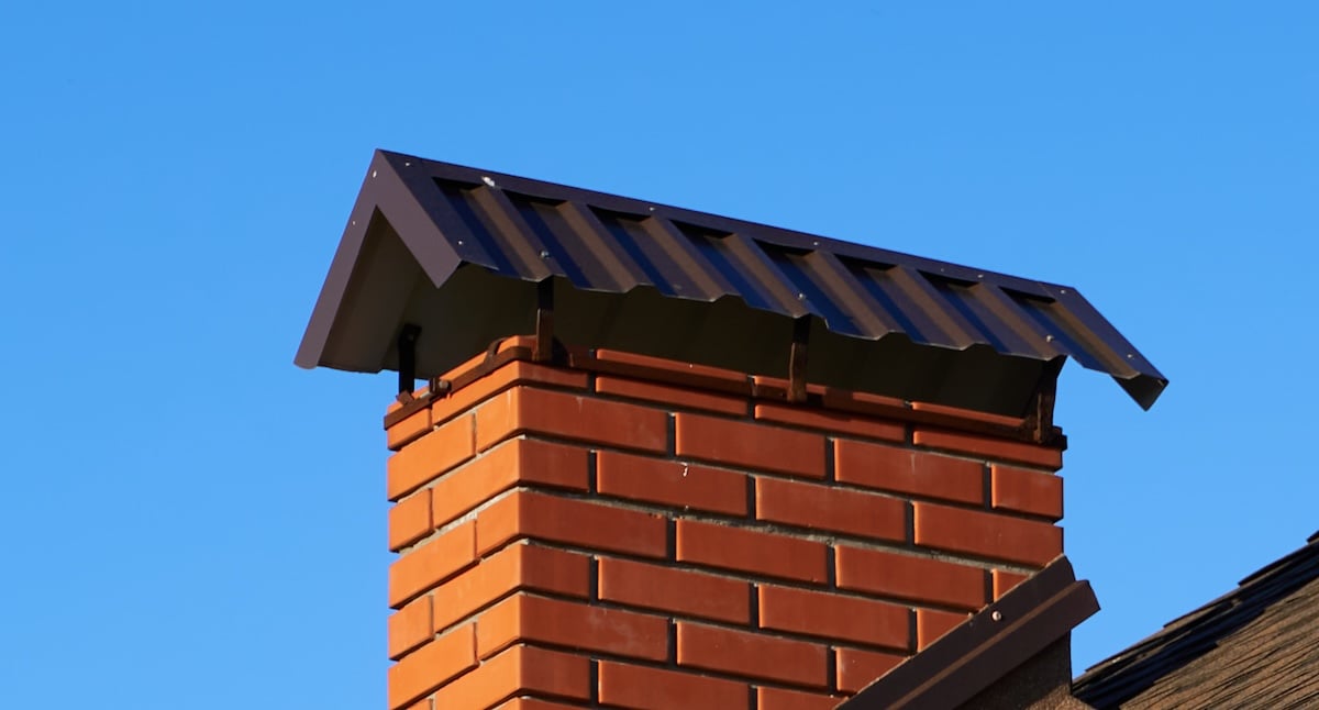 A close-up of a brick chimney with a dark metal cap, set against a clear blue sky. The metal cap has a sloped, ridged design and shades the top of the chimney.