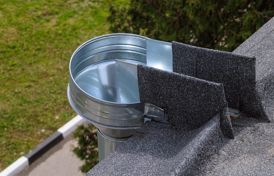 A round metal rain gutter on a roof directs water into a downspout. The gutter is bordered by two roof shingles and is set against grass and pavement in the background.